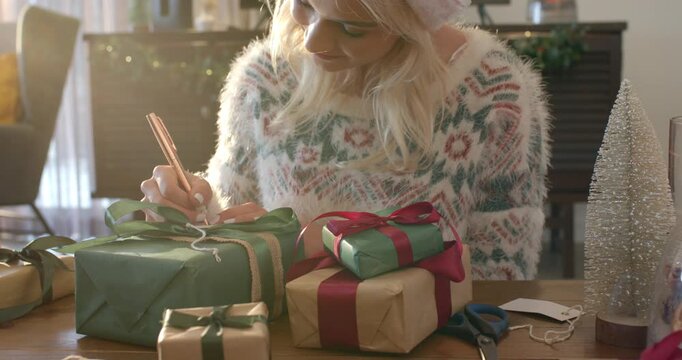 Picking up pen, woman is writing tag and tying bow on wrapped package for holiday gifts