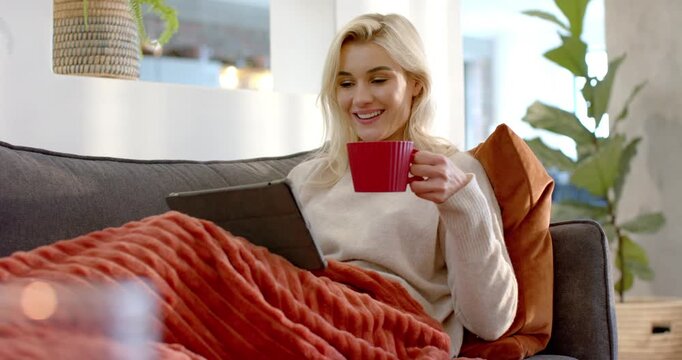 Adult woman noticing tablet headline tapping tablet and sipping red mug on charcoal sofa