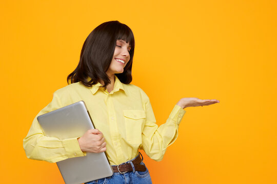 A cheerful woman in a yellow shirt smiles while holding a laptop against a vibrant orange background, creating a friendly, approachable office or study scene.