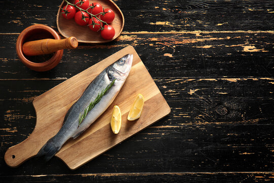 Cutting board with fresh raw seabass fish, lemon and cherry tomatoes on black wooden background - Powered by Adobe