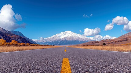 An empty asphalt road stretches towards a majestic snow-capped mountain range, framed by golden autumn trees and a bright blue sky.