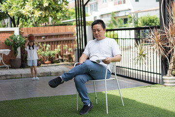 A man reads peacefully while a young girl holds a basketball in the background. A soft moment showing connection across generations at home.