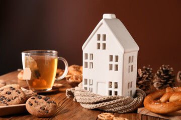 Winter composition with decorative house, forest bumps, cup of tea and tasty snacks on wooden table against brown background