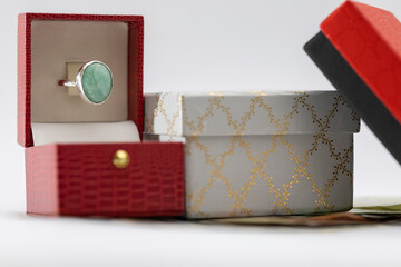 an elegant silver ring with a large aquamarine/emerald stone in an open red gift box, next to a decorative white and gold patterned box and a black box lid