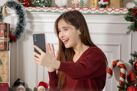 Asian young woman smiling and waving during christmas video call by decorated fireplace with gifts and candy cane