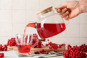 Woman pouring hot viburnum tea from glass teapot into cup on white tile background