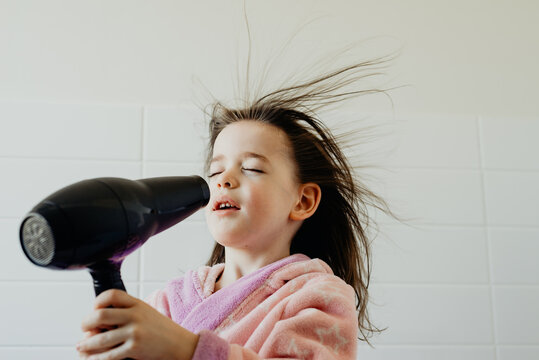 Portrait of a happy young girl wearing a bathrobe with her eyes closed blow drying her hair at home with a hair dryer in the bathroom, Australia