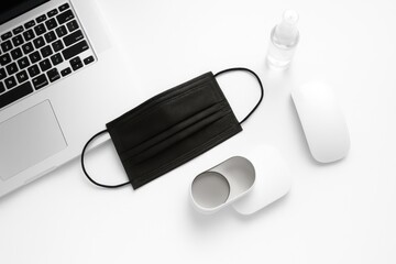 Black protective face mask with laptop mouse and hand sanitizer on clean white background office safety concept for covid remote work hygiene corporate health policy and workplace campaigns