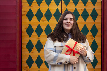 Street city photo. Young adult white woman with gift box with red bow stands near Santa Claus house with green rhombus pattern on yellow wooden wall at Merry Christmas fair. Happy New Year concept