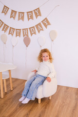 Adorable curly-haired girl sitting on white armchair in birthday party setup. Happy birthday banner with beige balloons against white wall and wooden floor.