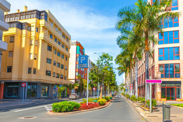 Sunlit street outside the Apartamentos Banosol in Las Palmas de Gran Canaria, typical urban scene with apartment buildings and palm trees lining road. Parking sign and manicured shrubs