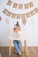 Happy little girl with blonde curly hair wearing a birthday hat, sitting at a white table in front of a party garland banner. Birthday celebration for child.
