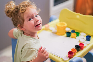 Young caucasian child with big eyes looking at camera. Curious toddler sitting at table with paints ready to draw or craft. Early childhood development and creative learning.