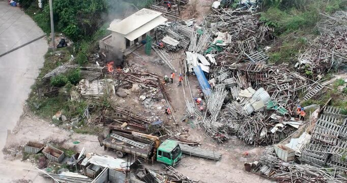 aerial view of a chaotic scrap yard reveals workers welding among massive metal piles, while a small fire burns near a white portable building.