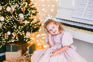 Little girl in a purple dress smiling at the camera as she sits next to a piano and a Christmas tree with glowing lights, for Christmas or New Year.