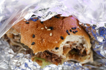 Close up of delicious pulled pork sandwich with sesame seed bun wrapped in foil. This satisfying, messy lunch food takeout meal with savory filling and fresh bread