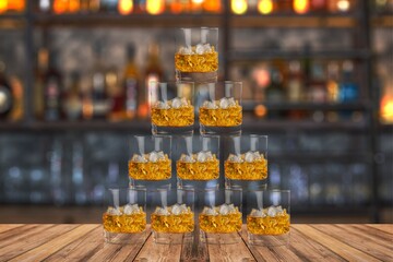 Pyramid of whiskey glasses filled with ice on a wooden bar surface, set against a blurred bar background, representing celebration and social drinking