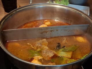 A metal hot pot with a divider containing two different boiling broths, showing meat, vegetables, and meatballs simmering in the savory liquid.