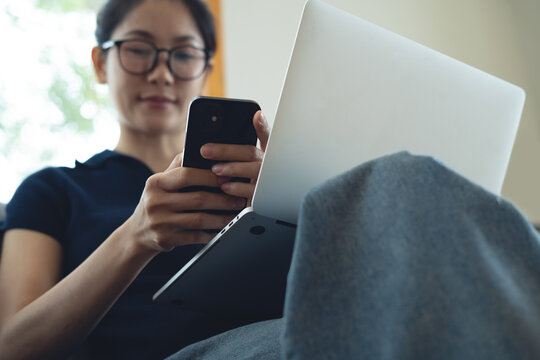 Woman using mobile phone and laptop while sitting on sofa at home. Young asian woman using smartphone searching in internet social network or remote working from home