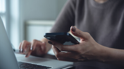 Woman using smartphone while working on laptop computer at home office, working remotely, freelance at work, online job, brown tone, closeup