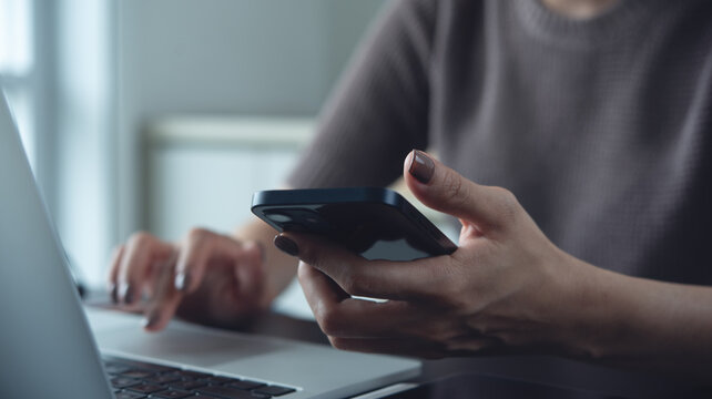 Woman using smartphone while working on laptop computer at home office, working remotely, freelance at work, online job, brown tone, closeup