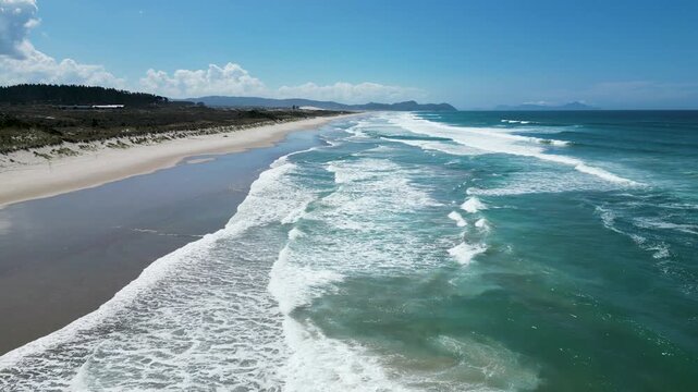 Cinematic low drone shot along Te Arai beach with rolling surf, white foam, and clean blue water. New Zealand.