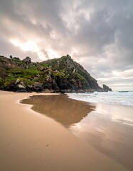 Obraz premium Coastal scene under a cloudy sky. A sandy beach reflects sunlight. A lush green, rocky hill rises from the water's edge, creating a dramatic landscape