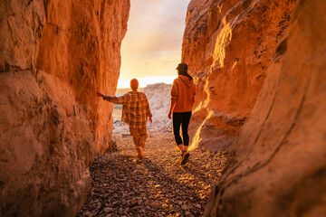 Two young women tourists are standing at lake or sea beach between big rocks at the exit from narrow canyon and looks at sunset shore