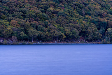 Smooth long-exposure water in soft light with dense autumn trees rising steeply from the shoreline of Llyn Padarn