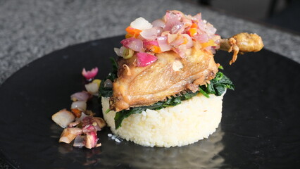 Plated Indonesian-style fried chicken topped with spicy sambal matah, served over leafy greens and steamed rice on black dish.
