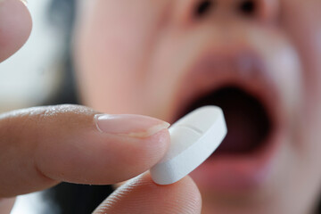 A person is about to take a pill, with their mouth open and the tablet held between their fingers,...