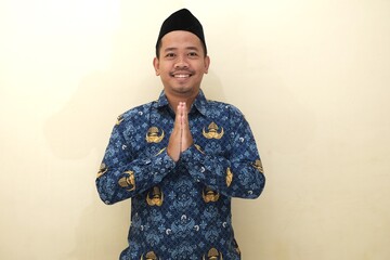 Smiling Indonesian man wearing civil servant (PNS) batik uniform and peci, greeting with traditional gesture, isolated with copy space for advertisement.
