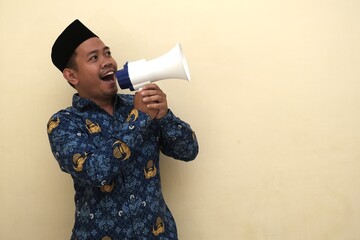 Energetic Indonesian man wearing civil servant PNS batik and peci shouting into a megaphone, isolated with copy space for advertisement or campaign.