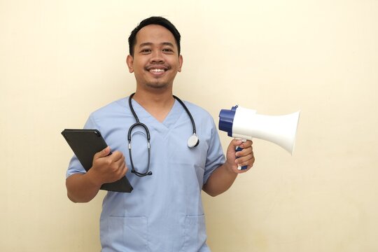 Smiling Southeast Asian man in scrubs with stethoscope holding tablet and megaphone, promoting health services or awareness; isolated with copy space for advertisement.