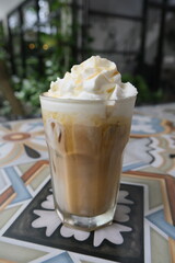 Refreshing iced coffee topped with whipped cream served in a glass with a straw, placed on a colorful patterned tile table outdoors.
