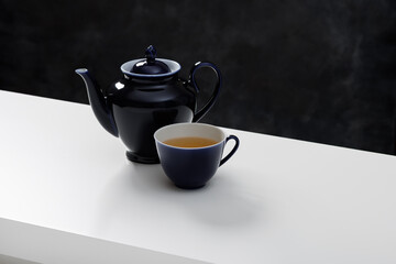 Fresh tea in a dark blue ceramic cup and a teapot on a white table. Dark background.
