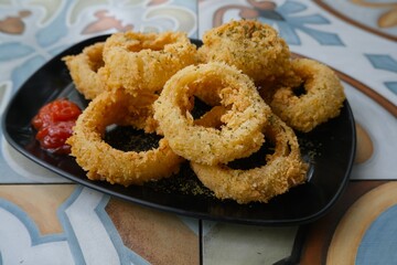 Golden brown onion rings served on a black plate, coated in crunchy breadcrumbs and sprinkled with herbs for a savory appetizer.