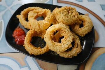 Golden brown onion rings served on a black plate, coated in crunchy breadcrumbs and sprinkled with herbs for a savory appetizer.
