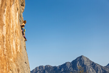 A strong girl climbs a rock against a backdrop of mountains and sky. Extreme hobby, Overcoming a difficult climbing route, Woman rock climbing. Effort during rock climbing,