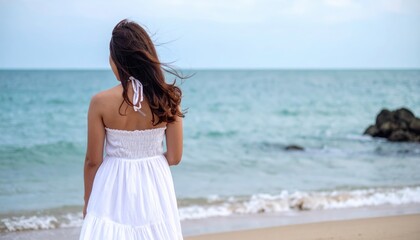 Young woman in white dress standing on beach, looking at ocean and enjoying tranquil seaside breeze during summer day