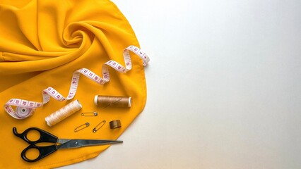 A composition with yellow fabric and a set of sewing tools, including thread, a measuring tape, scissors and a thimble with space for text.