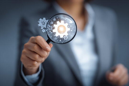 a businesswoman in a suit holds a magnifying glass with a light bulb and gears icon over her hand on a blurred office background, representing the concept of business ideas and innovative technology.