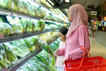 A Muslim mother in a pink hijab and her daughter shop for fresh vegetables in the supermarket. Healthy eating and family grocery shopping concept.