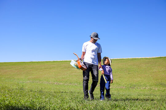 Father and His Daughter Walk Across a Sunlit Green Hill, Holding Hands and Carrying Their Picnic Gear