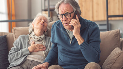 Mature man calling ambulance for his wife having heart attack on sofa at home
