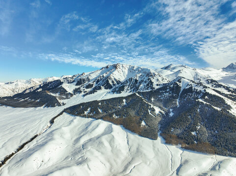 Expansive aerial view of rugged, snow-capped Tian Shan mountains and foothills meeting a wide, open snow-covered plain beneath a bright, dynamically cloudy blue sky