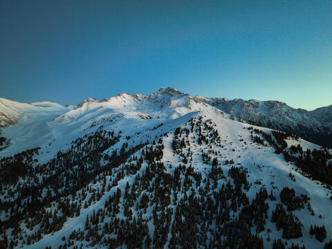 High angle aerial view of massive, sunlit, snow-capped mountain peaks and ridgelines dominating the skyline above the dark, snow-covered coniferous forest below at twilight