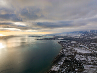 High angle aerial view of a snow dusted town on the coast of a large lake, with snowy mountains visible on the distant horizon under cloudy skies