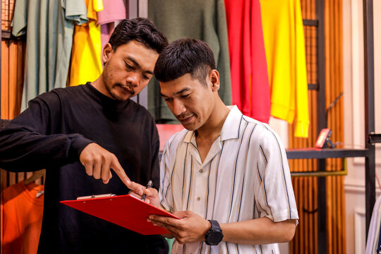 Two Men Collaborating on Clipboard Amidst Colorful Apparel in Modern Retail Setting