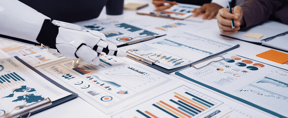 A modern business scene featuring a robotic hand assisting a professional team in analyzing charts, graphs, and reports on a collaborative workspace.Noogenesis